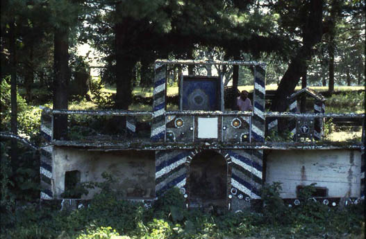 A small decorated grotto structure with blue-and-white striped framing in a wooded area