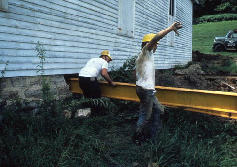 Two workers in yellow hard hats moving a yellow beam beside a white building