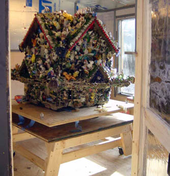 Decorated house-shaped folk-art structure covered in colorful trinkets on a wooden platform