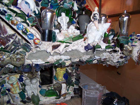 Mosaic-covered structure adorned with figurines, glass shards, and light bulbs