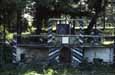 A small roofed structure and dark figure in front, set among trees and fence