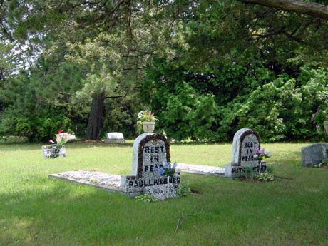 Flower-adorned gravestones in a grassy, tree-lined cemetery landscape