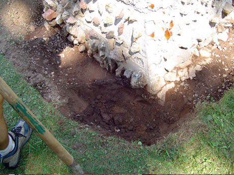 Dirt hole in grass beside a concrete wall and a wooden tool handle