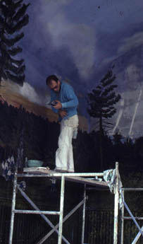 Man in blue shirt and white pants on scaffolding before painted forest backdrop