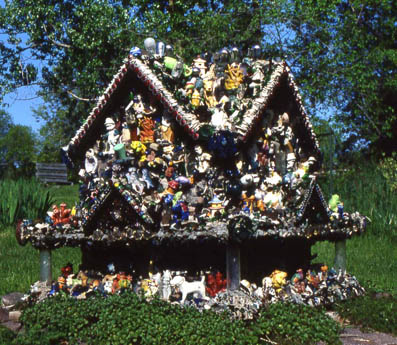 A small house-shaped structure densely covered with figurines and objects in a grassy yard