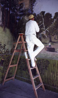 A figure in white on a ladder painting a forest mural on a wall