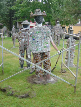Several bottle-cap-covered human figures standing on grass behind a metal fence